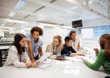 Teacher with a group of university students, in a laboratory classroom. The instructor is considering one of the students work, the mood is light hearted and positive. Other classmates are discussing things with each other. This is a realistic teaching scenario, with candid expressions. This is a multi-ethnic group of women. In the background there is a white board with mathematical formula written on it. All ladies are wearing id tags.