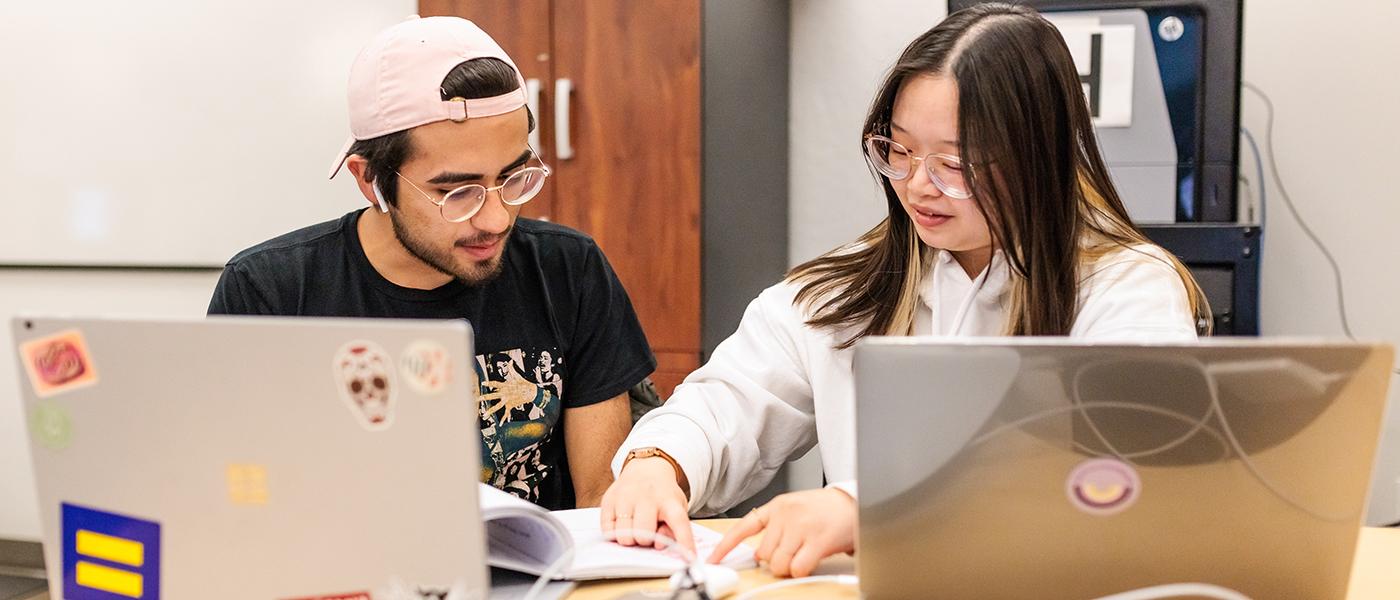 Two students sitting next to eachother to collaborate, each with thier own MacBook Pro laptop to work on.