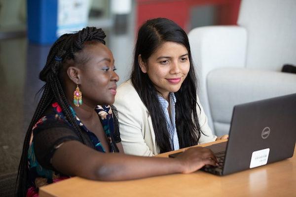 Two young woman sit side by side, looking at a laptop screen