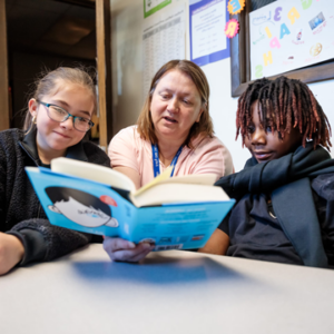 Photograph of teacher sitting at a table with two students reading a book.